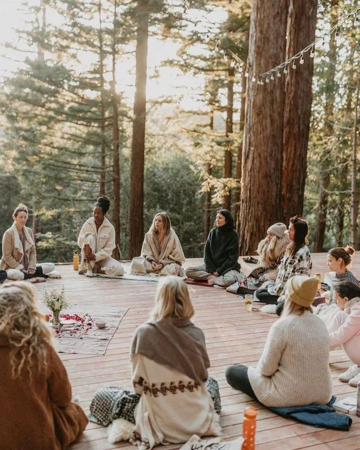 A circle of women sitting together on a wooden deck under tall trees, blankets over their shoulders and flowers laid in the centre